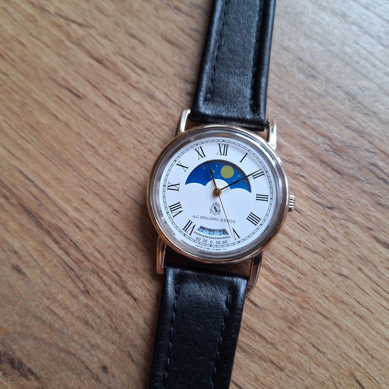 Op de afbeelding: Een goudkleurig polshorloge met een witte wijzerplaat met Romeinse cijfers en een maanfase-indicator. Het horloge heeft een zwarte leren band en de tekst "A.G. Spalding Bros." staat op de wijzerplaat.