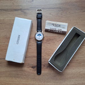 Op de afbeelding: Een Citizen quartz horloge met een zwarte leren band en een goudkleurige kast. De wijzerplaat van het horloge heeft een maanfase-ontwerp. Het horloge wordt getoond met de originele doos en documentatie, inclusief een "VEGA" merkkaart.