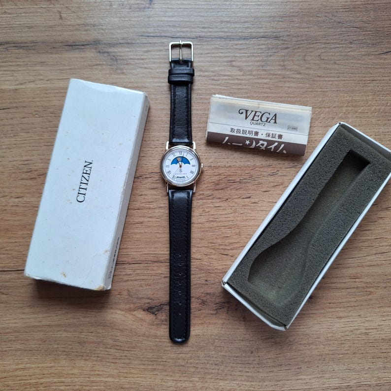 Op de afbeelding: Een Citizen quartz horloge met een zwarte leren band en een goudkleurige kast. De wijzerplaat van het horloge heeft een maanfase-ontwerp. Het horloge wordt getoond met de originele doos en documentatie, inclusief een "VEGA" merkkaart.