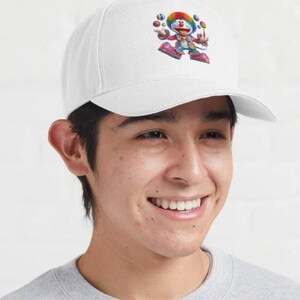 May include: White baseball cap featuring a colorful cartoon character juggling balls. The character has a rainbow-colored hat and is wearing a red and blue outfit. The cap is a classic baseball style with a curved brim.