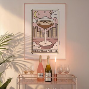 Puede incluir: Una ilustraci&oacute;n de carta de tarot con fondo rosa y una copa de espresso martini. La carta se titula "The Espresso Martini" y tiene el n&uacute;mero XII. La carta est&aacute; enmarcada y colgada en una pared.