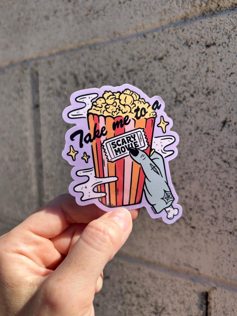 May include: A sticker featuring a cartoon popcorn bucket with the text "Take me to a Scary Movie." A zombie hand holds a movie ticket. The popcorn is yellow, and the bucket is red and orange.