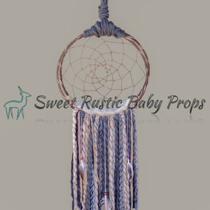 May include: A gray and white dream catcher with feathers hanging from the bottom. The dream catcher is hanging from a branch.
