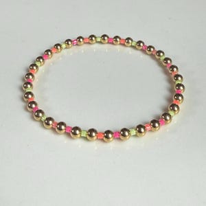 May include: Gold beaded bracelet with neon pink, orange, and green accents. The bracelet features small, round gold beads interspersed with colorful accent beads, creating a vibrant and playful design. This jewelry piece is suitable for everyday wear.