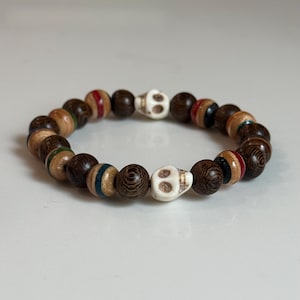Könnte beinhalten: Ein Armband aus dunkelbraunen Holzperlen, durchsetzt mit hellerem Holz und farbigen Akzenten in Rot, Blau und Grün. Zwei weiße Totenkopfperlen sind eingearbeitet. Das Armband ist ein stilvolles Accessoire.