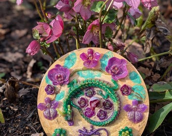 Puede incluir: Una piedra de jardín redonda y decorativa con un diseño de dragón verde y detalles morados, rodeada de flores y suculentas moradas. La piedra está colocada sobre un fondo de flores rosas y follaje.