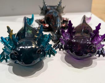 Puede incluir: Tres figuras de ajolotes coloridos. Los dos delanteros son de color azul oscuro y morado con branquias y volantes translúcidos. El tercer ajolote está en el fondo y es una mezcla de rojo oscuro y negro. Los tres tienen un acabado brillante.