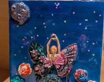 Puede incluir: Una pieza de arte de resina azul cuadrada con una bailarina con alas de mariposa. Las alas son rosas y verdes con detalles florales. El fondo tiene un diseño de noche estrellada con una luna y flores coloridas.