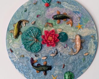 Puede incluir: Arte mural redondo que representa una escena de estanque con peces koi, nenúfares y una flor de loto roja. La obra de arte tiene un fondo marmoleado azul y blanco con varios elementos decorativos.