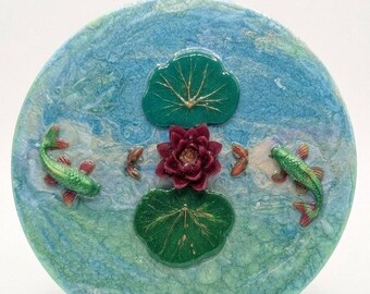 Puede incluir: Una pieza decorativa redonda con un diseño acuático en tonos azules y verdes. Presenta una flor roja, hojas de nenúfar verdes y peces verdes con detalles rojos. El texto "Whisper of Peace: Awakening Flow" está en la parte superior.