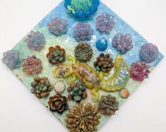 Puede incluir: Una pieza de arte mural en forma de diamante con un lagarto amarillo rodeado de varias figuras de suculentas de resina en tonos azul, morado, verde y marrón. Una placa dorada en la parte inferior muestra el texto, THE DESERT QUEEN'S FLORAL DREAM.