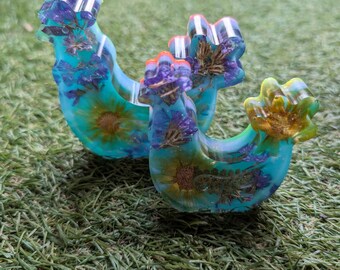 Puede incluir: Dos objetos decorativos de resina curvados, cada uno con una base turquesa y flores secas incrustadas. Las partes superiores son una mezcla de morado, amarillo y naranja. Los objetos están sobre una superficie verde.