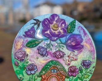 Puede incluir: Placa decorativa redonda con un diseño de jardín de hadas. La placa tiene una combinación de colores verde y morado con una casa de hadas, flores y una mariposa. También aparece un pájaro negro en la placa.
