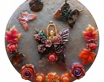 Puede incluir: Una pieza decorativa redonda y gris con una superficie texturizada, adornada con coloridas mariposas, flores y hojas. Una figura central con los brazos extendidos está rodeada de elementos florales. La pieza presenta una variedad de colores, incluyendo rojo, naranja y morado.