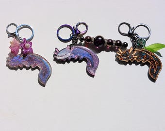 Puede incluir: Tres llaveros con forma de ajolote en tonos morado, azul y dorado. Cada llavero tiene un diseño diferente, uno con flores rosas y perlas, otro con cuentas oscuras y el último con una hoja verde.