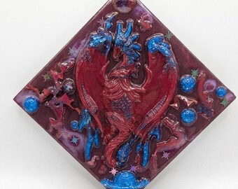 Puede incluir: Una obra de arte de resina en forma de diamante que presenta una criatura mítica roja y azul con alas extendidas. La obra incluye elementos celestiales como estrellas y planetas, con el texto "Guardians of the Starlit Sky" en la parte inferior.