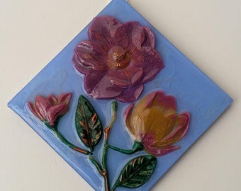 Puede incluir: Escultura floral en relieve con una flor grande, texturizada, de color púrpura oscuro con un centro dorado, dos capullos y hojas verdes sobre un fondo azul en forma de diamante. La obra de arte está montada en una pared blanca.