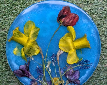 Puede incluir: Un posavasos redondo de resina azul con flores secas incrustadas. El posavasos presenta narcisos amarillos, flores silvestres moradas y amarillas y una flor de color rojo oscuro. Las flores están dispuestas sobre un fondo azul.