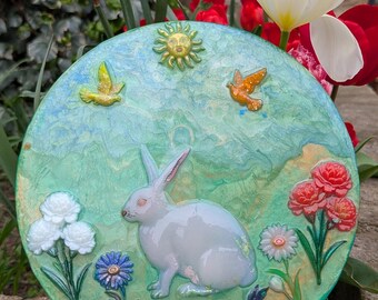 Puede incluir: Placa decorativa redonda de cerámica con un conejo blanco, flores y pájaros sobre un fondo verde y azul. La placa tiene una superficie texturizada y se exhibe sobre un soporte transparente. El sol también está representado.