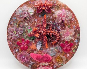 Puede incluir: Una pieza de arte de resina redonda con una figura central de dragón y adornos florales y de mariposas circundantes. La pieza presenta una mezcla de tonos rosas, rojos y naranjas, con el texto "Reborn Glow Dragon" en la parte inferior.