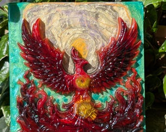 Puede incluir: Una obra de arte cuadrada de resina que representa un fénix rojo que se eleva de las llamas. La obra de arte tiene un fondo turquesa y dorado, y se exhibe en un caballete negro. El fénix tiene las alas extendidas y un centro amarillo.