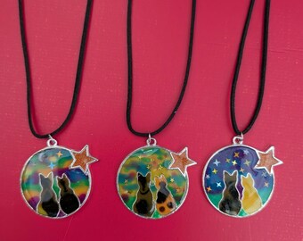 Puede incluir: Tres colgantes redondos en cordones negros, cada uno con dos siluetas de gatos sobre un fondo de cielo nocturno colorido con una pequeña estrella brillante a un lado. Los diseños muestran gatos mirando las estrellas en varios tonos de azul, verde y naranja.
