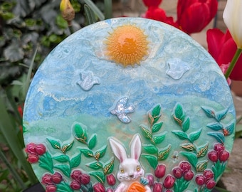 Puede incluir: Un plato decorativo redondo de cerámica con una escena de jardín primaveral. El plato tiene un cielo azul con un sol amarillo y nubes blancas. Un conejo blanco sosteniendo una zanahoria está rodeado de flores rojas y follaje verde.