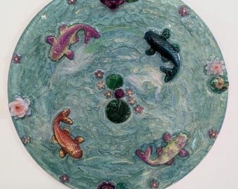 Puede incluir: Plato decorativo redondo de color turquesa con un diseño de estanque de carpas koi. El plato tiene cuatro coloridas carpas koi, nenúfares y detalles florales. El plato tiene aproximadamente 30 cm de diámetro.