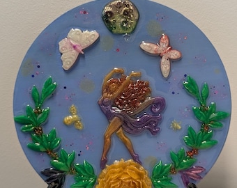 Puede incluir: Un plato decorativo redondo azul con una figura danzante, mariposas y una luna. El plato presenta una flor amarilla en la parte inferior y detalles de hojas verdes. El plato se exhibe sobre un soporte negro.