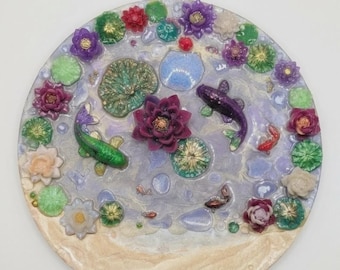 Puede incluir: Una pieza decorativa redonda con un diseño de jardín acuático. Presenta flores moradas, rojas y blancas, nenúfares verdes y peces koi. El texto "Whisper of Peace: Harmony in Bloom" está en la parte superior y "May your home always have a corner of peace" en la inferior.