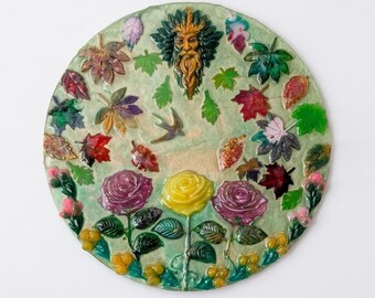 Puede incluir: Una placa decorativa circular titulada Emerald Grove Spirit que presenta un rostro verde central rodeado de hojas de otoño coloridas, un pájaro pequeño, dos rosas moradas, una rosa amarilla y bayas amarillas sobre un fondo verde claro.