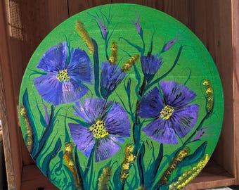 Puede incluir: Una obra de arte redonda pintada a mano con vibrantes flores moradas con centros amarillos sobre un fondo verde brillante. La pintura incluye tallos verdes y detalles amarillos, montada en un pequeño caballete de madera. La obra se exhibe en una caja de madera.