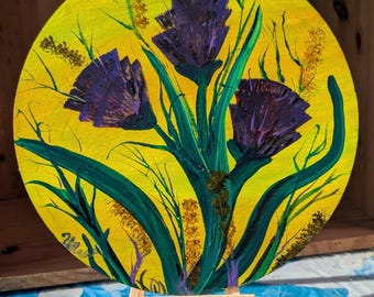 Puede incluir: Pintura redonda de flores moradas con tallos y hojas verdes sobre un fondo amarillo. La obra de arte se exhibe en un pequeño caballete de madera. La pintura tiene una apariencia texturizada, con pinceladas visibles.