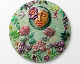 Puede incluir: Una placa de pared circular en verde claro con un rostro central de tono dorado rodeado de flores esculpidas en tonos rosa, morado y rojo, con pequeños pájaros y hojas. Debajo de la placa hay una placa de color latón con la inscripción The Flora Mystique.