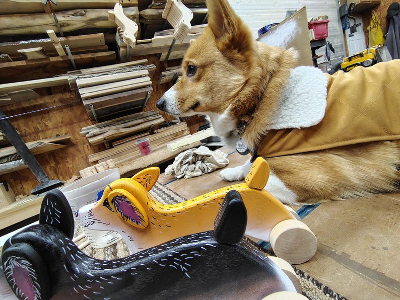May include: Two wooden toy dogs, one black and white and one yellow, with wheels. A brown and white dog is looking at the toys.