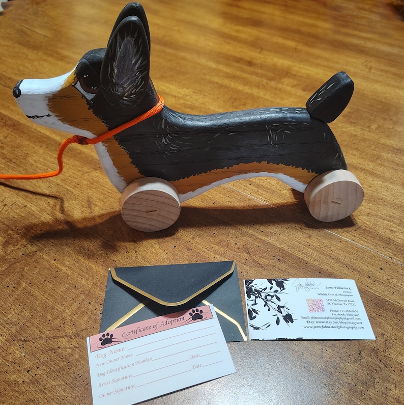 May include: A wooden toy dog painted in black, brown, and white with orange leash. The dog has four wheels and is a pull toy. The toy is accompanied by a certificate of adoption and a business card.
