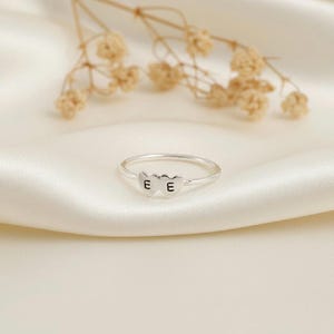 May include: A silver ring with two heart-shaped charms, each bearing the letter "E". The ring rests on a white satin surface, with dried flowers in the background. The ring is a delicate piece of jewelry.
