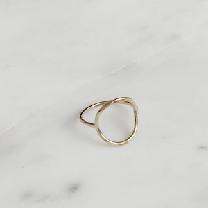 Puede incluir: Un anillo dorado con un diseño de círculo abierto. El anillo tiene una textura martillada y una banda delicada. El anillo está sobre una superficie de mármol blanco.