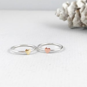 May include: Two silver rings with a small heart-shaped accent. One ring has a gold heart and the other has a rose gold heart.