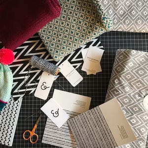 May include: A flat lay of various fabrics and packaging materials for gift wrapping. The fabrics include a teal and white geometric pattern, a gray and white geometric pattern, and a black and white geometric pattern. There are also several white cards with the text "&" printed on them. The text "Ravel & Unravel" is printed on a white card.