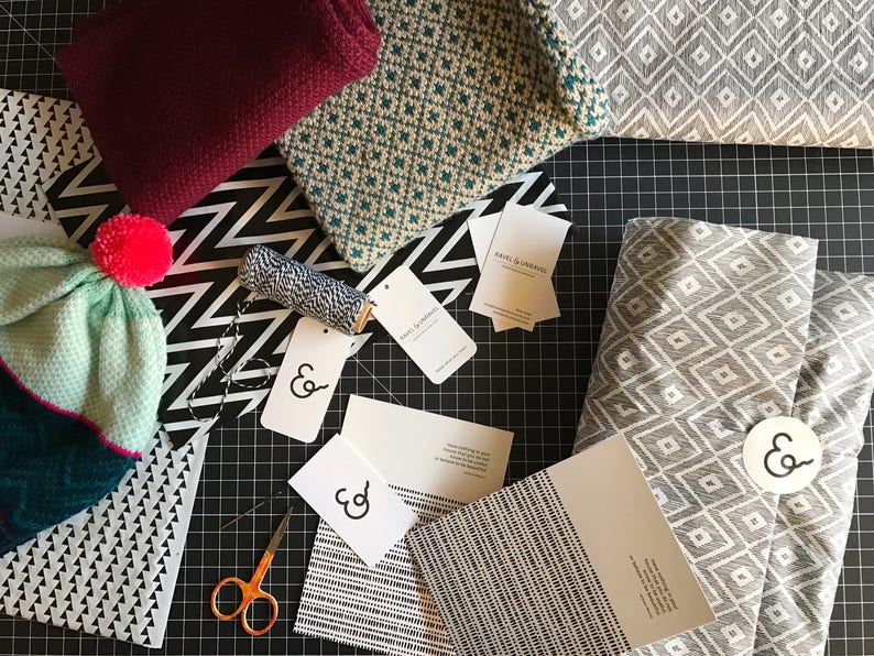 May include: A flat lay of various fabrics and packaging materials for gift wrapping. The fabrics include a teal and white geometric pattern, a gray and white geometric pattern, and a black and white geometric pattern. There are also several white cards with the text "&" printed on them. The text "Ravel & Unravel" is printed on a white card.