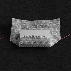 May include: A white paper package with a geometric pattern and a label that reads "RAVEL & UNRAVEL". The package is tied with a pink string.