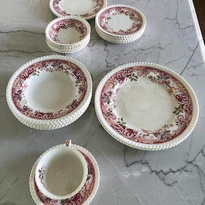 May include: A collection of white ceramic dishware with a floral pattern in shades of burgundy, pink, and green. The set includes plates, bowls, and a teacup with a matching saucer. The edges of the plates and bowls have a textured, rope-like design.