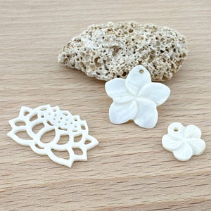 Może przedstawiać: Trzy białe muszelkowe zawieszki w kształcie kwiatów, dwa to kwiaty frangipani, a jeden ma bardziej skomplikowany wzór. Zawieszki leżą na jasnym drewnianym podłożu.