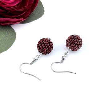 Pode incluir: Um par de brincos pendentes com contas esféricas cobertas por pequenas sementes de cor vermelho-escura. Cada conta está presa a um gancho de metal prateado com uma pequena tampa decorativa na base da esfera, criando uma textura.