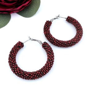 Pode incluir: Um par de brincos de argola com um design circular coberto por pequenas contas vermelho-escuras. Os brincos têm fechos de metal prateado e estão posicionados perto de uma grande flor vermelho-escura com folhas verdes num fundo branco.