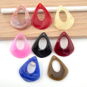 Op de afbeelding: Een verzameling van acht druppelvormige acryl sieradenhangers met een hol midden en een gladde, gemarmerde afwerking. De hangers zijn verkrijgbaar in verschillende kleuren, waaronder roze, rood, crème, zwart, bordeauxrood, blauw en bruin.