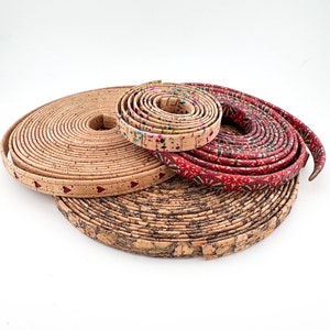 Puede incluir: Varias tiras de corcho enrolladas con diferentes diseños. Una tira es de corcho natural con corazones rojos, otra tiene un patrón moteado colorido y una tercera es roja con un diseño floral blanco. Sobre un fondo blanco.