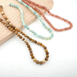 Puede incluir: Tres hilos de cuentas de piedras preciosas en varios colores: marrón, azul claro y rosa. Las cuentas son redondas y están ensartadas en un cordón blanco. Las cuentas marrones tienen un aspecto moteado. Las cuentas se muestran sobre una superficie de madera clara.