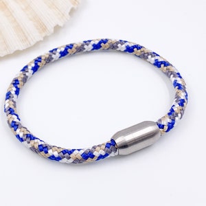 Puede incluir: Un brazalete de cuerda trenzada azul, blanco y beige con un cierre magn&eacute;tico plateado.